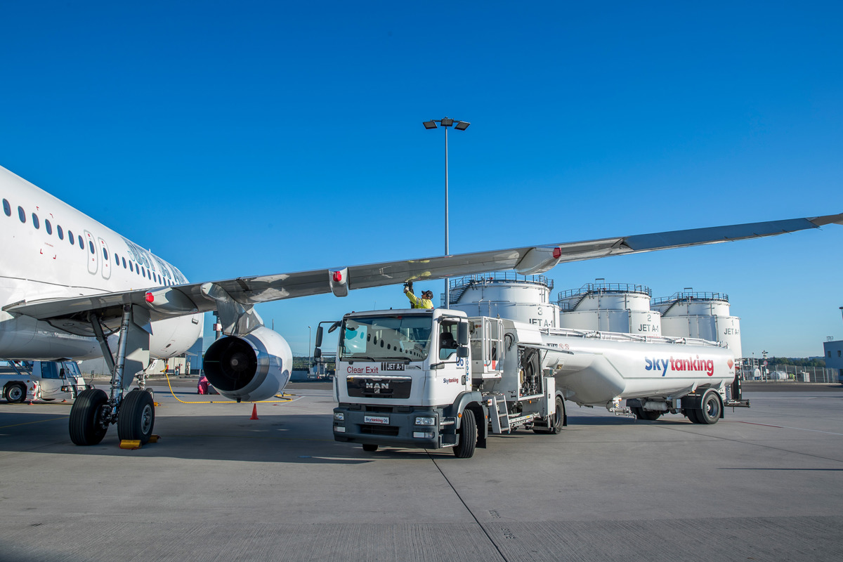 Flugzeug wird von Tankfahrzeug betankt, im Hintergrund das Tanklager