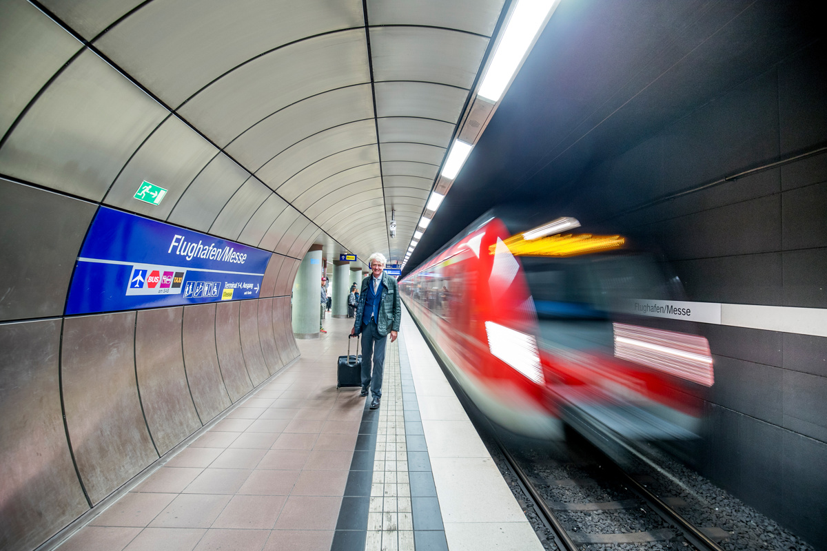 S-Bahn fährt an der Haltestellt Flughafen/Messe ein und Mann mit Koffer läuft auf dem Bahnsteig